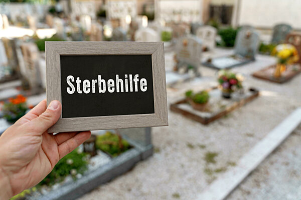 22 August 2023: Hand Holding A Plaque With The Text Euthanasia In A Cemetery With Gravestones PHOTOMONTAGE
