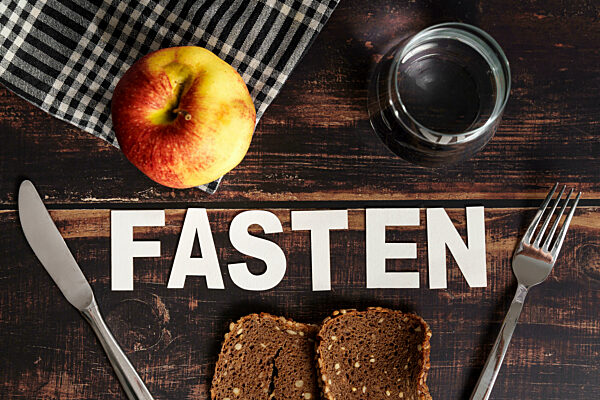 Schrifzug Fasten auf einem Holztisch neben altem Brot, einem Glas Wasser und einem Apfel. Symbolbild Fastenzeit
