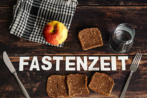 Schrifzug Fastenzeit auf einem Holztisch neben altem Brot, einem Glas Wasser und einem Apfel. Symbolbild Fastenzeit von Aschermittwoch bis Ostern