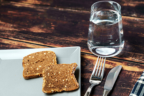 Fastenzeit Symbolbild, ein Glas Wasser neben einem Teller mit trockenen Brot