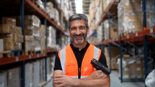 Logistikmanager mittleren Alters, der mit verschränkten Armen in einem großen Gang des Distributionszentrums steht, einen Barcodescanner in der Hand hält und in die Kamera lächelt. Er zeigt Selbstvertrauen und Fachwissen