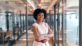 Professionelle junge Geschäftsfrau mit Afro Frisur, die selbstbewusst lächelt, während sie mit verschränkten Armen im Flur eines modernen Büros steht und Stolz und Erfolg zeigt