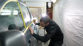 Fleißiger Mechaniker bereitet ein Fahrzeug für eine neue Lackierung vor, indem er in einer professionellen, für die Lackierung ausgestatteten Karosseriewerkstatt vorsichtig Klebeband auf den Autotürfensterrahmen aufträgt