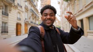 Junger schwarzer männlicher Vlogger, der in einer bezaubernden Pariser Straße ein Selfie Video für soziale Medien filmt, lächelt und zeigt begeistert auf den ikonischen Eiffelturm im Hintergrund