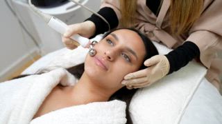 Entspannte junge Frau, die von einer professionellen Kosmetikerin eine nichtinvasive Mikrostrom Gesichtsbehandlung erhält, um die Muskeln zu stimulieren, den Hautton zu verbessern und Falten in einem modernen Spa zu reduzieren