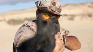 Ein männlicher Soldat, der in Wüstentarnung gekleidet ist, konzentriert sich intensiv, während er sein Gewehr mit durchsichtiger Schutzbrille in einer sandigen Umgebung richtet