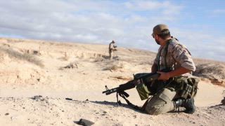 Zwei männliche Militärsoldaten beim Kampftraining in einer Wüste, einer kniete mit einem Gewehr, während ein anderer Mann eine Position im Hintergrund einnimmt