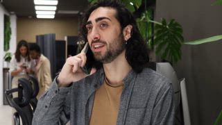 Ein Mann beim Telefonieren in einem modernen Büroraum, umgeben von viel Grün und modernem Dekor. Hemd mit Kragen, Stuhl, Mitarbeiter im Hintergrund, die ein aktives Arbeitsumfeld veranschaulichen