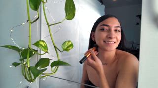 Eine Frau lächelt, während sie sich mit einem Pinsel schminkt, was sich in einem Spiegel neben grünen Ranken widerspiegelt und Schönheit und natürliche Elemente in Harmonie hervorhebt