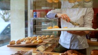 Professioneller Bäcker in weißer Uniform, der sorgfältig frisch gebackenes Gebäck und Croissants auf einer hölzernen Vitrine in einer gemütlichen Backstube, vom Schaufenster aus gesehen, arrangiert