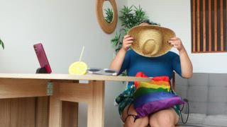 Transmann im Urlaub, entspannt sich mit Regenbogentasche, Sonnenhut und Zitronengetränk. Symbol für Stolz und Selbstentfaltung in einer gemütlichen, stilvollen Umgebung