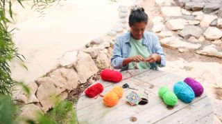 Eine Frau sitzt draußen an einem runden Holztisch, umgeben von lebendigen Garnen, und bastelt. Die ruhige natürliche Umgebung verleiht ihrer kreativen Routine ein friedliches Ambiente