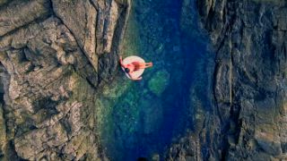 Blick von oben auf eine nicht wiederzuerkennende Frau, die sich auf einem aufblasbaren Boot in einem ruhigen, schmalen Naturpool, umgeben von schroffen Klippen, am Strand von Tindaya auf Fuerteventura entspannt