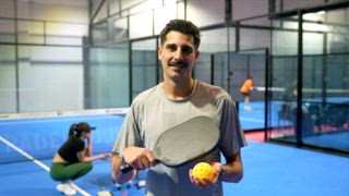 Lächelnder männlicher Athlet mit Schnurrbart, der ein Pickleball Paddel und einen gelben Ball auf einem blauen Hallenplatz hält und in die Kamera schaut, während andere Spieler im verschwommenen Hintergrund trainieren