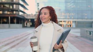 Eine Frau gemischter Abstammung mit lockigem Haar genießt einen Moment in der Stadt und hält eine Kaffeetasse und eine Tablette in der Hand. Warm gekleidet verkörpert sie den urbanen Lebensstil und die moderne Arbeitskultur