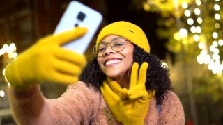 Fröhliche junge Frau in gelber Winterkleidung, die in einer weihnachtlich geschmückten Innenstadt Selfies mit ihrem Smartphone macht
