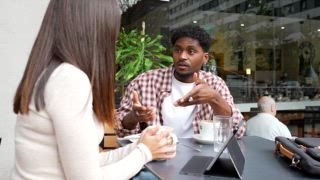 Junge gemischtrassige Freunde sitzen an einem Cafétisch im Freien, wobei der Mann begeistert gestikuliert, während er mit der Frau spricht, die aufmerksam zuhört, während sie eine Kaffeetasse in der Hand hält