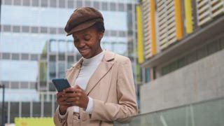 Eine urbane schwarze Frau lächelt, während sie in der Stadt ihr Handy checkt. Sie trägt einen beigen Mantel und eine Mütze, die einen modernen und stilvollen Look verkörpern. Die Szene fängt urbane Eleganz und Konnektivität ein