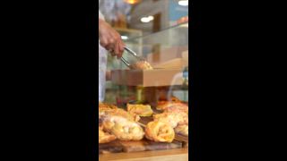 Ein Bäckereiangestellter legt mit einer Zange eine frische, warme Empanada für einen Kunden in eine Pappschachtel zum Mitnehmen. Auf einem Holzbrett hinter der Glastheke wird eine Vielzahl köstlicher Backwaren ausgestellt