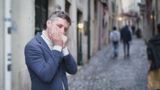 Ein Mann spielt Mundharmonika, während er in einer engen Straße in Lissabon, Portugal, steht. Die Leute gehen vorbei, während er auftritt und sein musikalisches Talent in der Stadt unter Beweis stellt