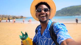 Junger schwarzer Mann mit Rucksack, Hut und Sonnenbrille genießt den Sommerurlaub und macht Selfies am Strand