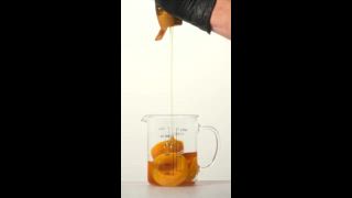 Eine Hand mit schwarzen Handschuhen drückt eine Honigflasche zusammen und träufelt goldenen Honig über geschnittene Pfirsiche in einem durchsichtigen Glasmessbecher. Der saubere weiße Hintergrund unterstreicht die reichen Texturen und Farben