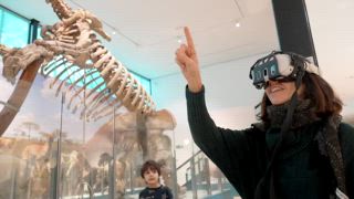 Lächelnde Frau mit Virtual Reality Headset und gestikuliert mit ihren Händen. Erkundung einer interaktiven Ausstellung mit einem riesigen Faultierskelett in einem Naturkundemuseum. Ein immersives Erlebnis schaffen