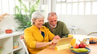 Glückliches älteres Paar, das zu Hause ein digitales Tablet benutzt, auf den Bildschirm zeigt und lächelt, während es zusammen frühstückt