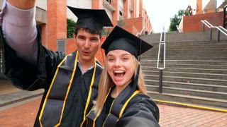 Fröhliches junges Paar in Abschlusskleidern und Mützen macht ein Selfie Video, lächelt und zeigt ihre Diplome in die Kamera, während sie ihre akademischen Leistungen an der Universität feiert