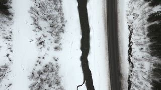 Dieses mit einer Drohne aufgenommene Video zeigt eine nordische Winterszene in Norwegen mit einem gefrorenen Fluss, der parallel zu einer Asphaltstraße verläuft und inmitten verschneiter Landschaften einen wunderschönen Kontrast bildet