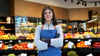 Porträt einer schönen jungen Arbeiterin, die mit verschränkten Armen in die Kamera lächelt und stolz vor einer Obst und Gemüseabteilung in einem modernen Lebensmittelgeschäft steht