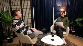 Junger Mann und Frau sitzen in Sesseln, unterhalten sich und nehmen einen Live Stream Podcast oder ein Interview auf, während ein Freund im Hintergrund tanzt und dann das Set verlässt