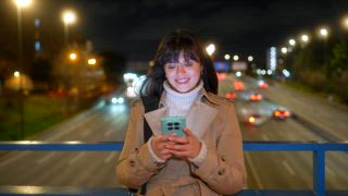 Lächelnde junge Frau, die glücklich ihr Smartphone benutzt, während sie nachts auf einer Brücke über einer stark befahrenen Autobahn steht. Der helle Bildschirm beleuchtet ihr Gesicht und die Autolichter erzeugen einen Bokeh Effekt