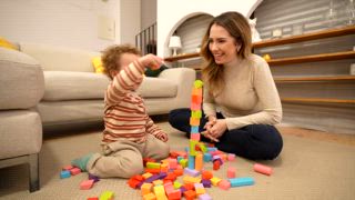 Lächelnde Mutter spielt mit ihrem kleinen Sohn und baut einen hohen Turm mit bunten Holzklötzen auf den Boden des Wohnzimmers, bis er zusammenbricht und sie zusammen lachen
