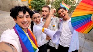 Eine bunt gemischte Gruppe lächelnder Freunde, die während einer Pride Parade ein Video für die sozialen Medien aufnimmt, eine Regenbogenfahne schwenkt und ein Megaphon in der Hand hält, um die Rechte und die Freiheit der Homosexuellen mit Freude zu feiern