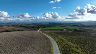 Ein malerischer Blick auf eine kurvenreiche Straße durch die sanften Hügel der Marken in Italien. Grüne Felder, verstreute Häuser und dramatische Wolken schaffen eine ruhige ländliche Landschaft