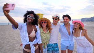 Verschiedene Freunde, die unvergessliche Momente festhalten, lachen und Selfies machen, während sie einen sonnigen Tag am Mittelmeerstrand genießen, und so eine unbeschwerte Sommerfreundschaft zeigen