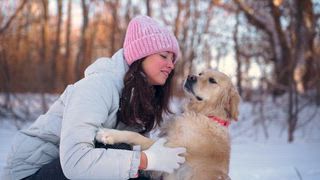 Eine fröhliche Frau umarmt ihren Labrador Retriever herzlich, während sie im Schnee spielt. Ihre Nähe und die winterliche Landschaft sorgen für einen freudigen und liebevollen Moment im Freien