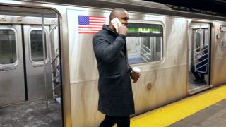 Afroamerikanischer Geschäftsmann mit einer Kaffeetasse verlässt eine U Bahn, während er auf seinem Smartphone spricht. Er lächelt, als er in einer belebten U Bahnstation in New York City den Bahnsteig entlang geht