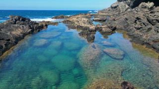 Eine Drohnenaufnahme aus der Luft zeigt eine nicht wiederzuerkennende Frau, die sich an den natürlichen Schwimmbädern von Aguas Verdes auf Fuerteventura auf den Kanarischen Inseln entspannt. Das zerklüftete Gelände umgibt klares türkisfarbenes Wasser