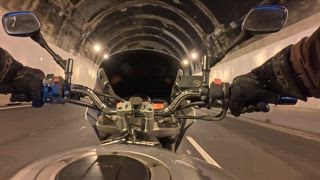 Ansicht eines Motorradfahrers aus der ersten Person, der auf einer Asphaltstraße fährt und in einen dunklen, beleuchteten Tunnel einfährt. Dabei sind Lenker, Spiegel und Armaturenbrett mit wechselndem Licht zu sehen
