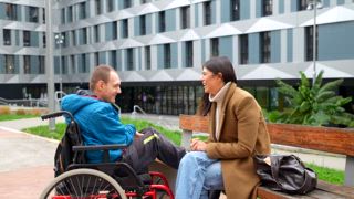 Junger Mann mit einer körperlichen Behinderung, der im Rollstuhl sitzt und mit seiner weiblichen Bezugsperson eine Therapiesitzung im Freien durchführt. Er spricht und lächelt zusammen auf einer Bank vor einem modernen Gebäude