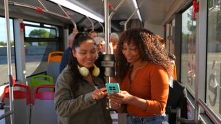 Zwei unterschiedliche junge Frauen, die lächeln und Spaß miteinander haben, während sie während ihres täglichen Pendelns in einem öffentlichen Bus ein Selfie mit einem Smartphone machen und auf Reisen einen glücklichen Moment teilen
