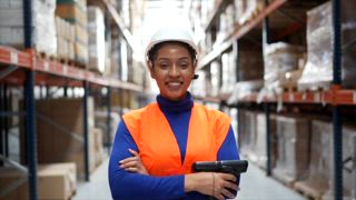 Porträt einer stolzen afroamerikanischen Arbeiterin mit Barcodescanner, die in einem großen Logistiklager steht und zwischen Regalen aus Pappkartons selbstbewusst in die Kamera lächelt