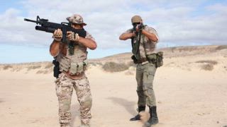 Militärs in Wüstenlandschaft, ausgerüstet mit Gewehren und Schutzausrüstung, trainieren in Schießstellung inmitten von sandigem Gelände