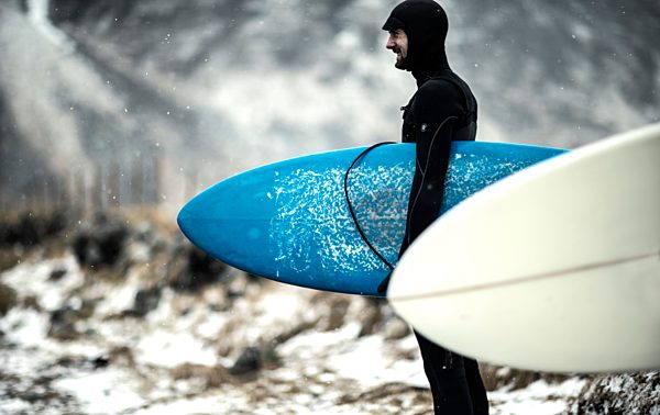 A surfer wearing a wetsuit and carrying surfboards standing on a snowy beach with mountains behind.