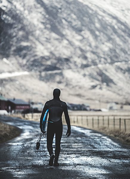 Rear view of a surfer walking on a road wearing a wetsuit and carrying a surfboard.