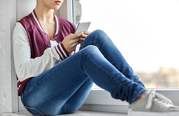 teenage girl with smartphone sitting on windowsill