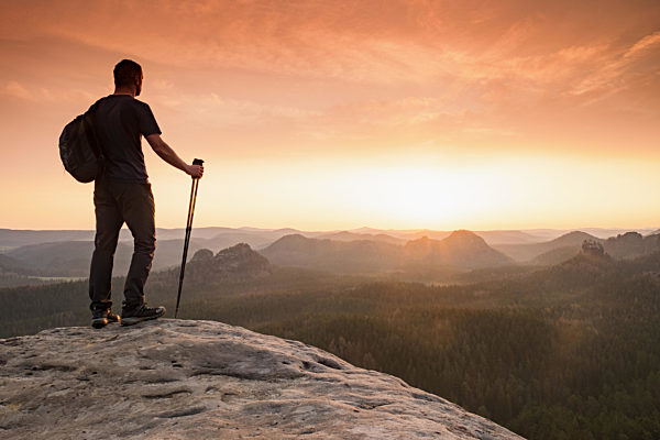 Hiker with backpack leaning on trekking poles staring into misty sunrise.