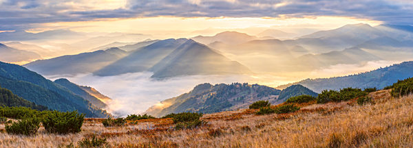Autumn morning mountain panoramic view with sunbeams through haze and low clouds.